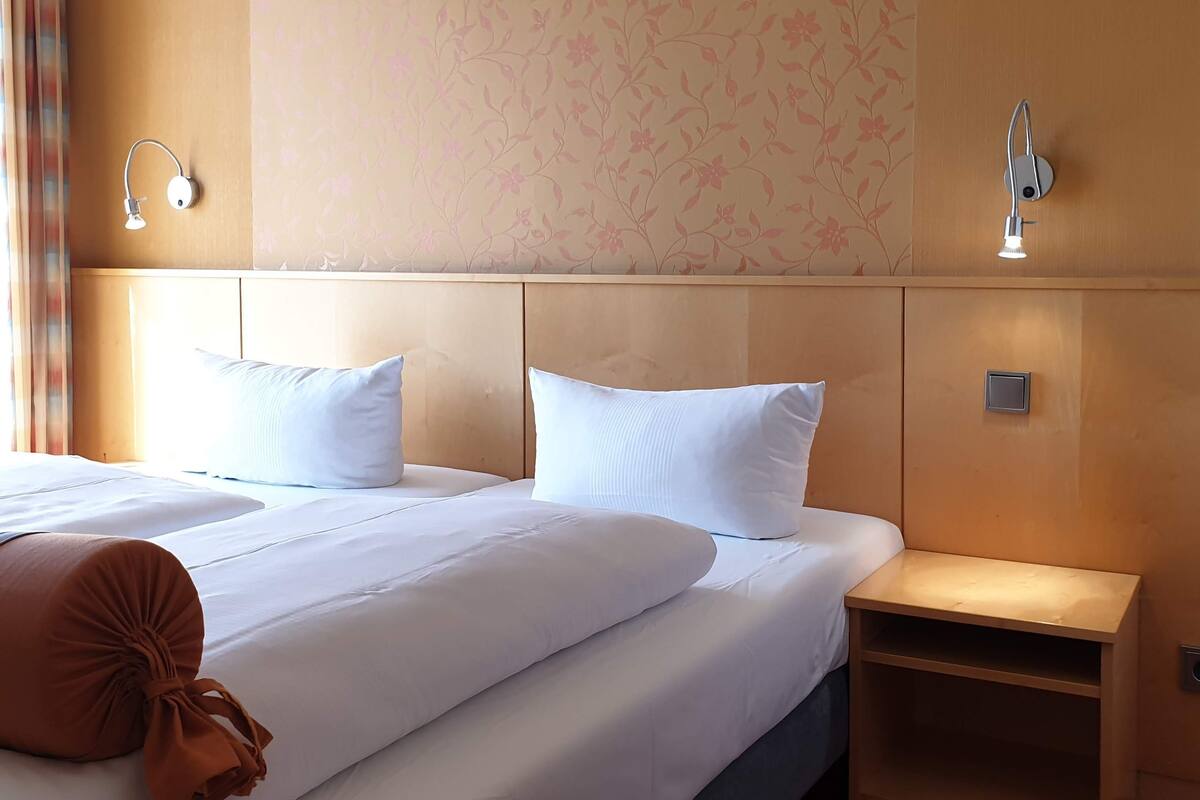 A hotel room with a bed, two pillows, a bedside table, and a wall-mounted light fixture.