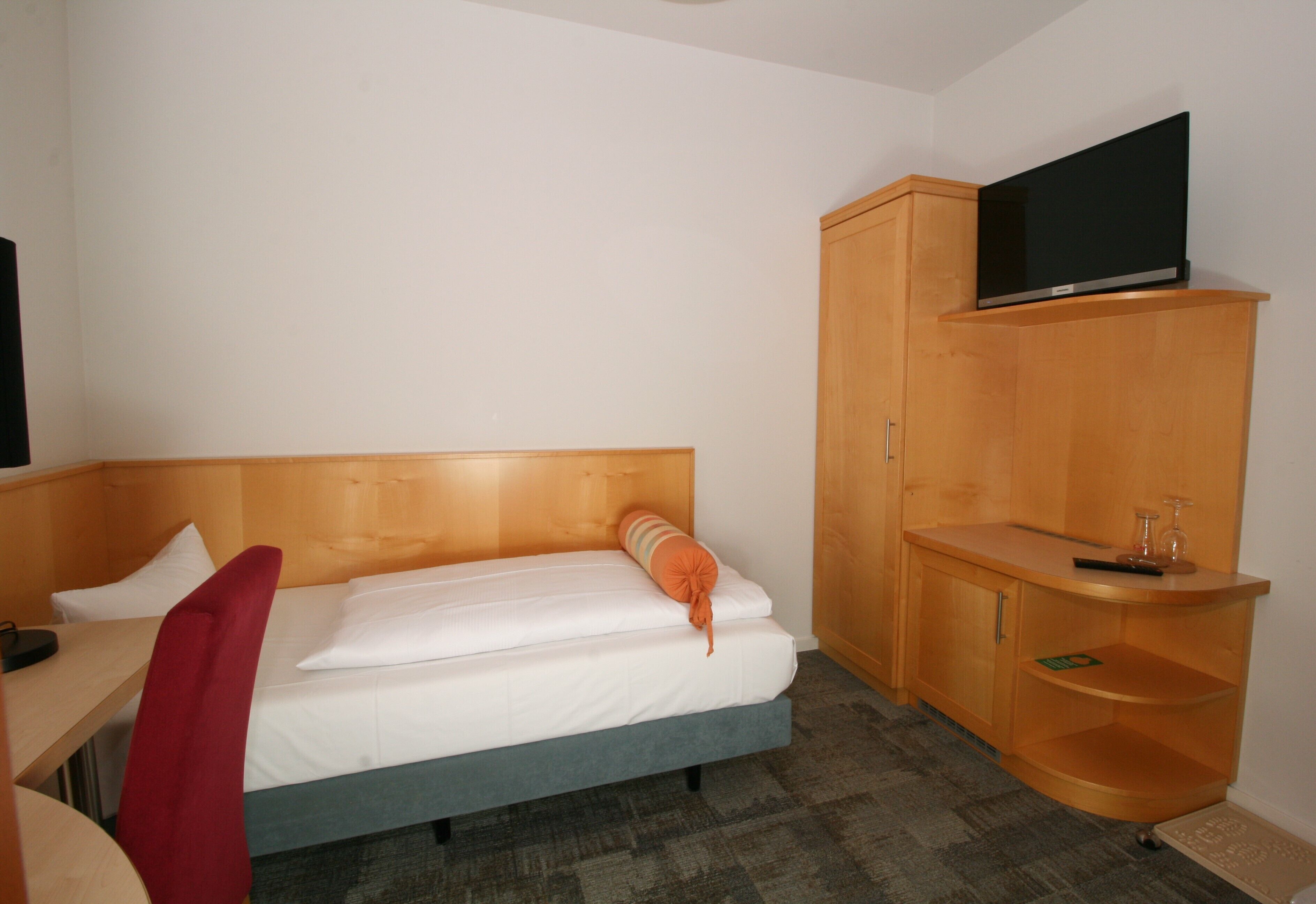 A hotel room with a bed, a desk, a TV mounted on a wooden cabinet, and a small shelf with a remote control.