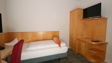A hotel room with a bed, a desk, a TV mounted on a wooden cabinet, and a small shelf with a remote control.