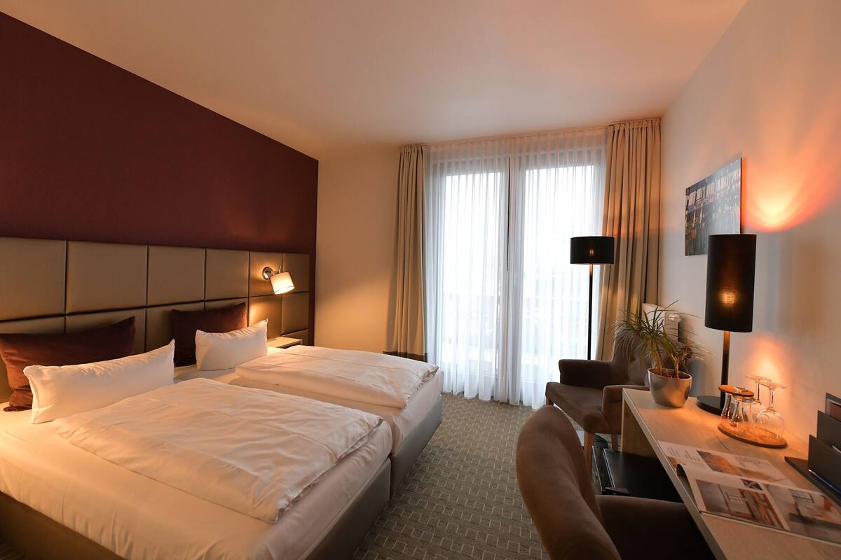 A hotel room with two beds, a desk, a chair, a potted plant, and a large window with curtains.