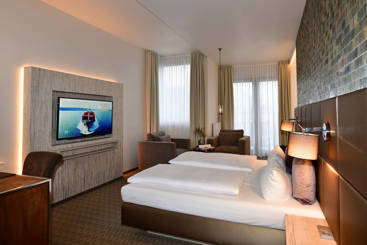 A hotel room with a large bed, a desk, a television mounted on the wall, and a view of the sea through the windows.
