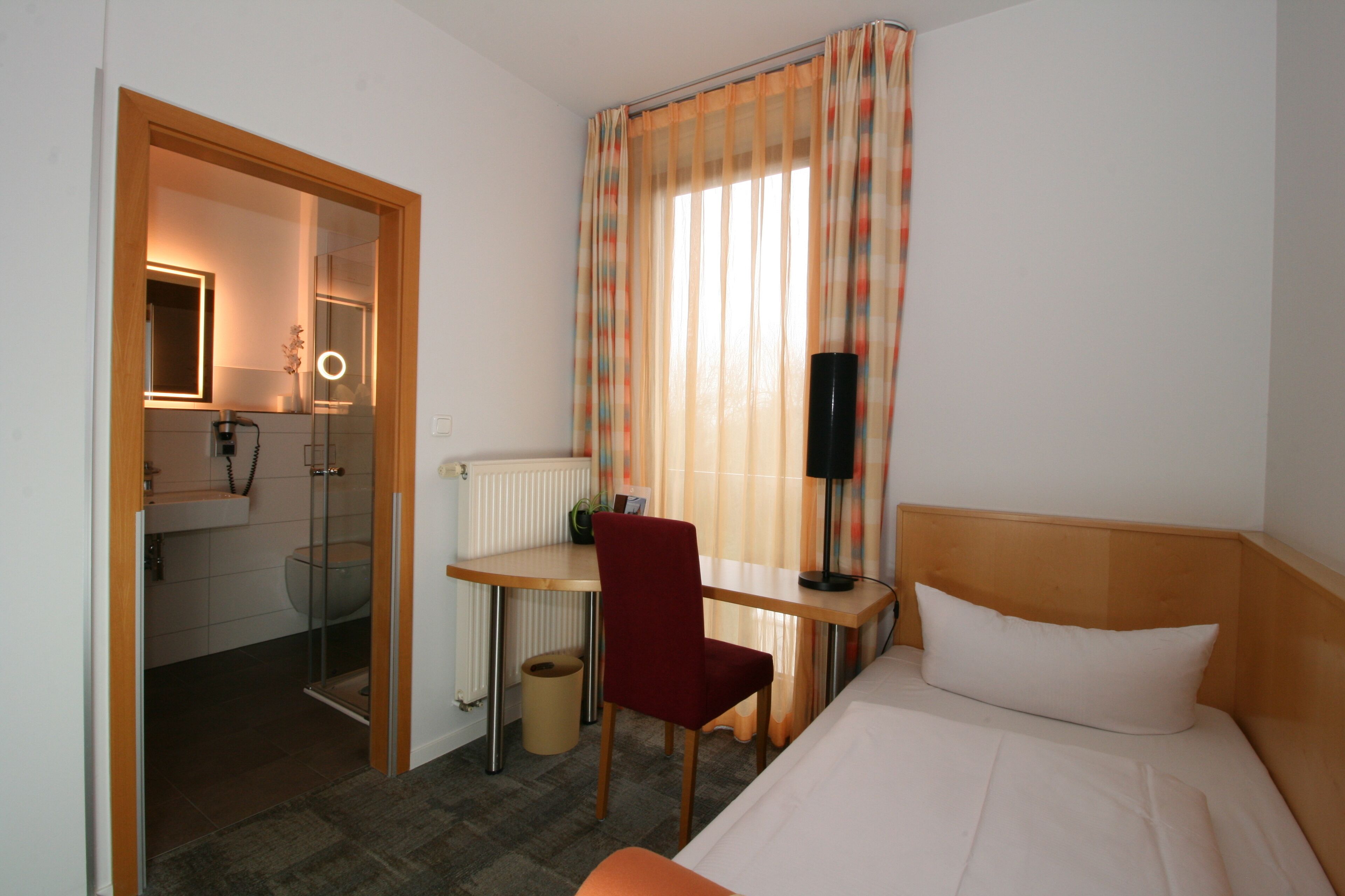 Ein Hotelzimmer mit einem Bett, einem Schreibtisch mit Stuhl, einem Spiegel, einem Fenster mit Vorhängen und einem durch eine offene Tür einsehbaren Badezimmer.