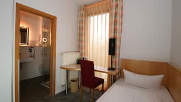 Ein Hotelzimmer mit einem Bett, einem Schreibtisch mit Stuhl, einem Spiegel, einem Fenster mit Vorhängen und einem durch eine offene Tür einsehbaren Badezimmer.