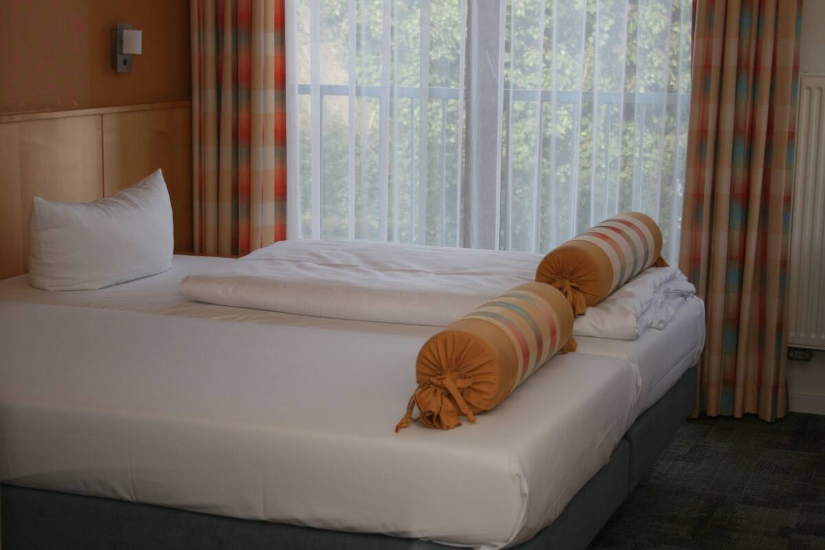 A double bed with white linens and striped pillows, a headboard, and a window with curtains.