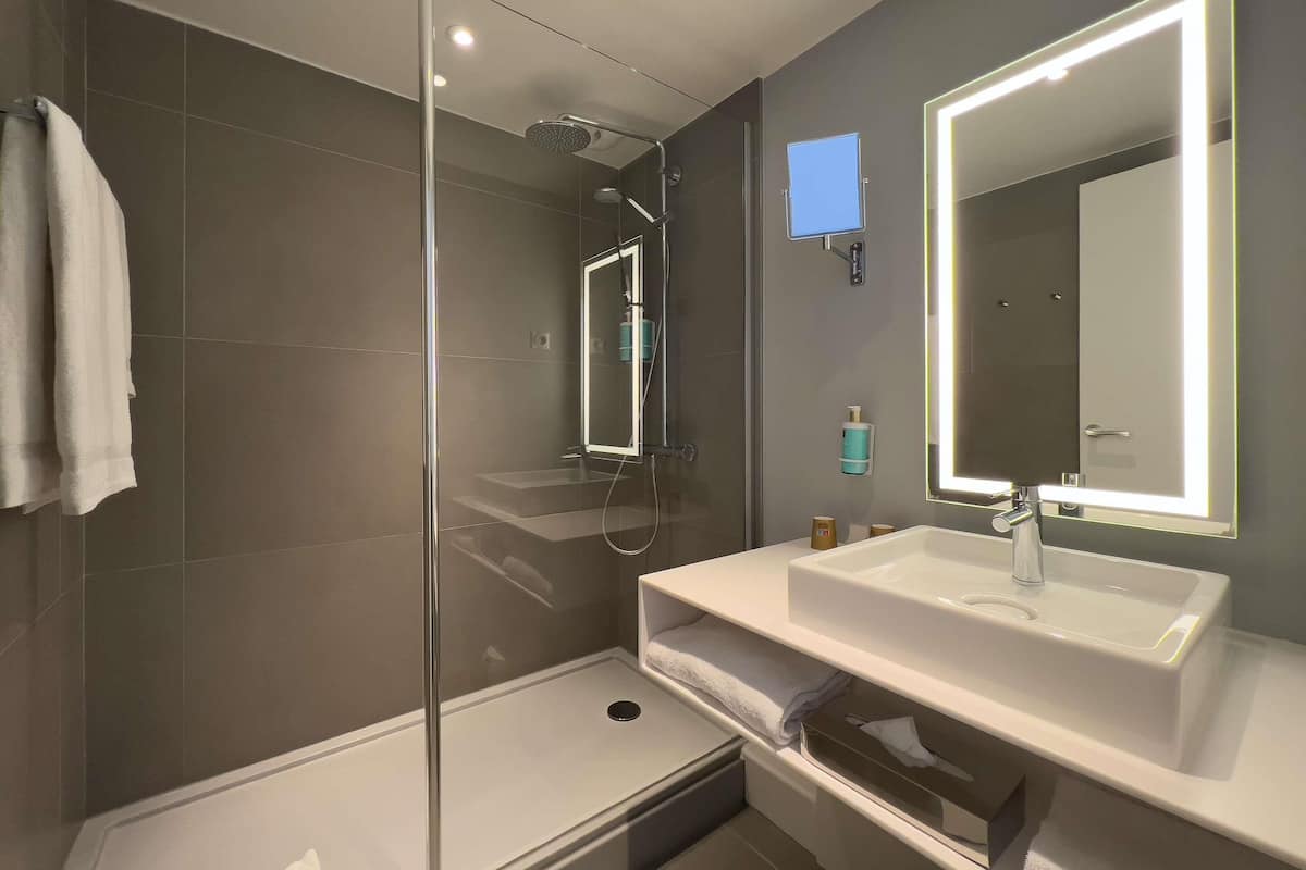 Un baño moderno con ducha envuelta en vidrio, un espejo grande con iluminación indirecta y un lavamanos blanco.