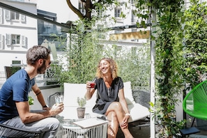 Guests can enjoy a peaceful conversation in this serene outdoor seating area, surrounded by lush greenery and a charming white building.