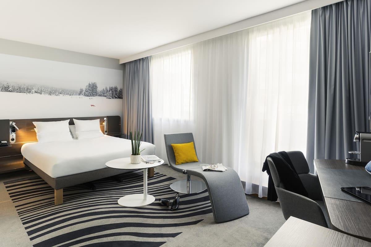 Habitación de hotel moderna con una cama grande, un escritorio, una silla y vistas a un paisaje nevado a través de cortinas translúcidas.
