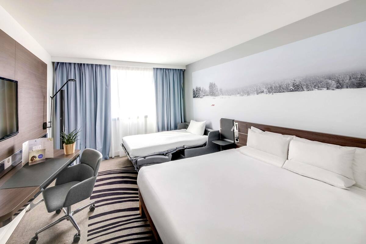Una habitación de hotel con una cama, dos mesitas de noche, un escritorio con una silla, un televisor y un ventanal con cortinas.