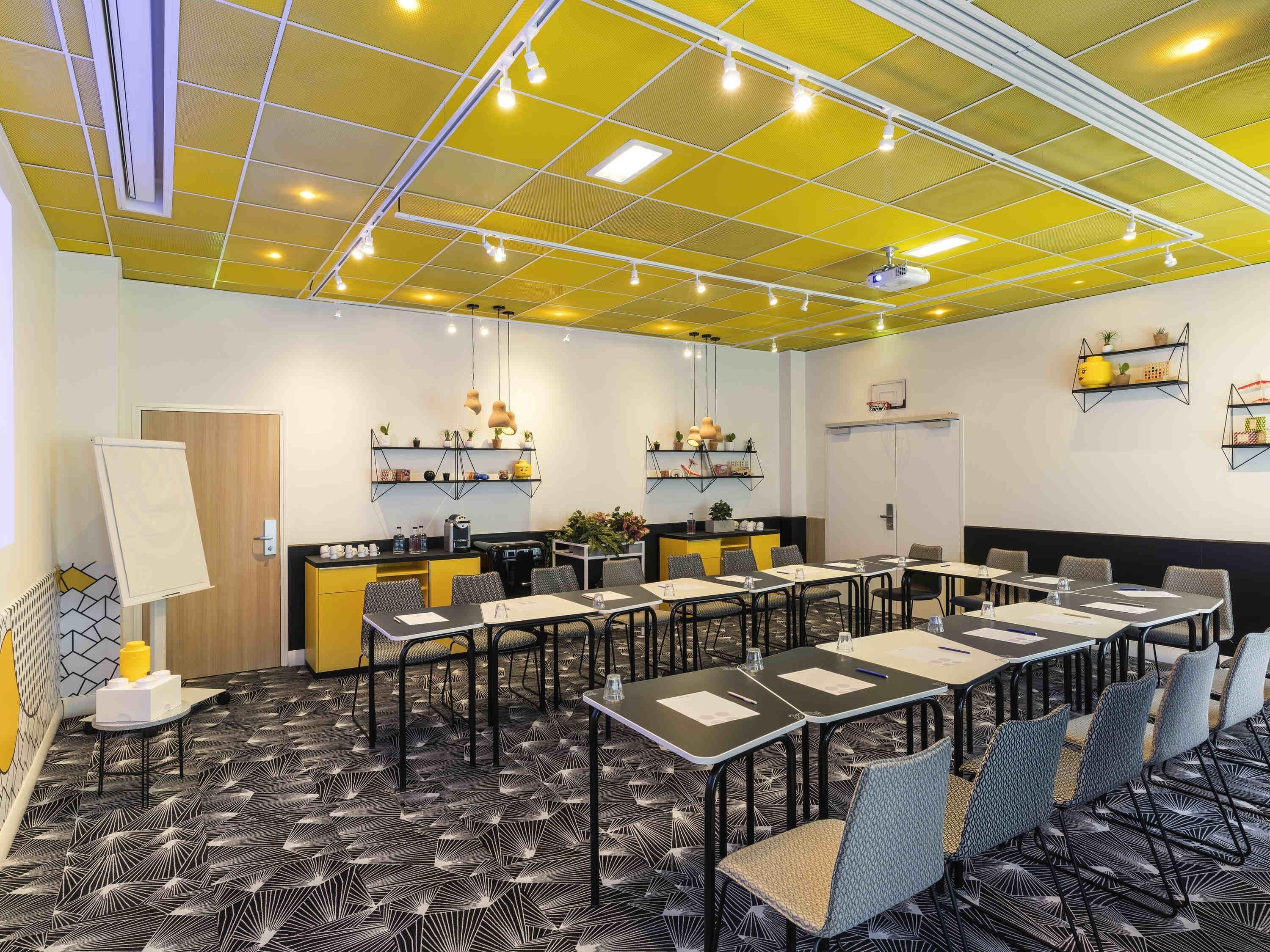 Sala de conferência com mesas, cadeiras, quadro branco e estantes com plantas.