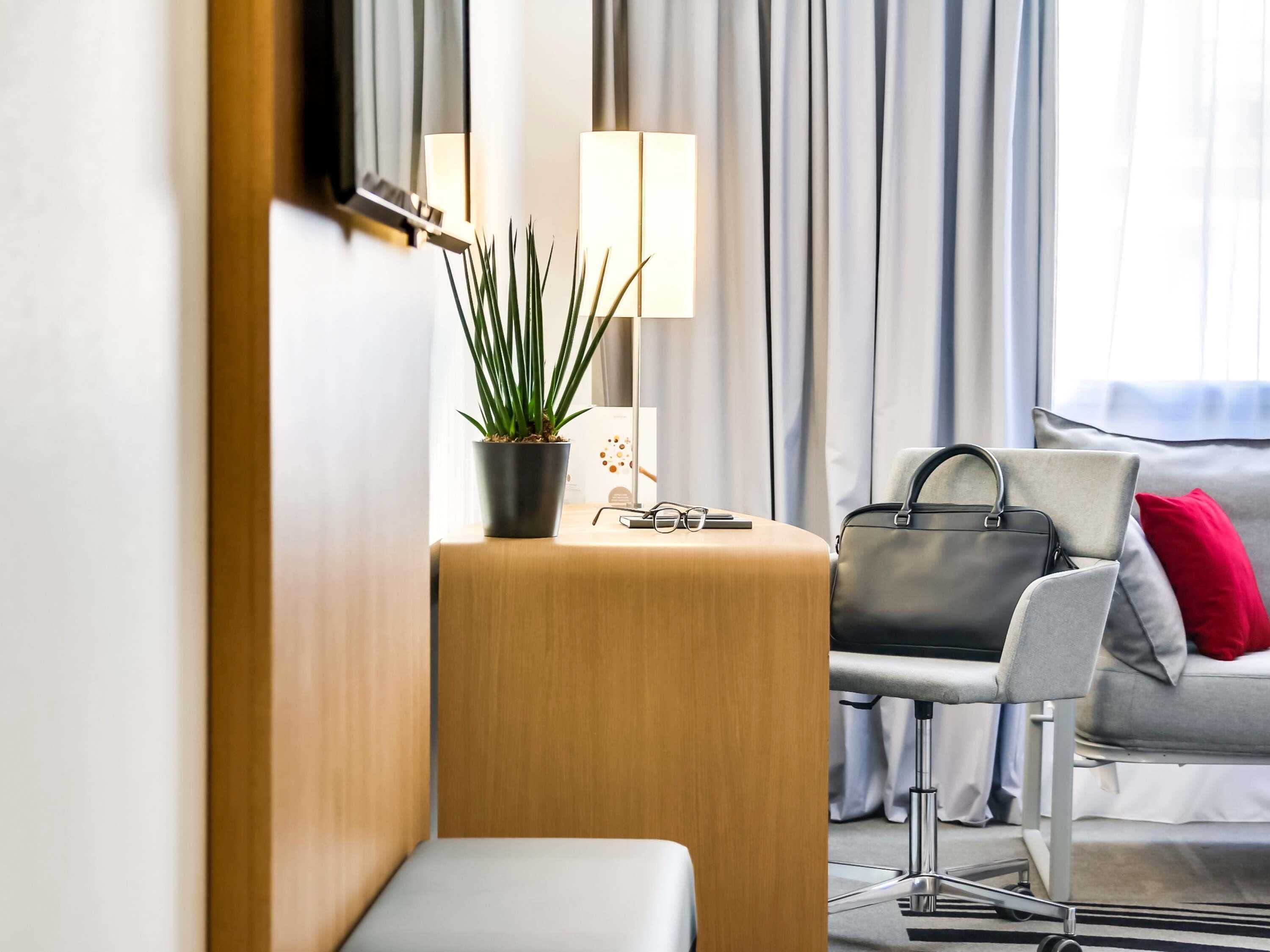 A hotel room with a wooden sideboard, a potted plant, a desk with a lamp, a grey armchair with a red pillow, and a bed with grey bedding.