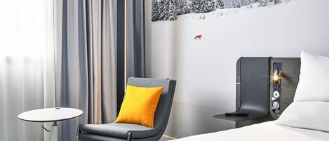 Una habitación de hotel moderna con una cama, una silla con un cojín amarillo, una mesita y una mesita de noche con un control remoto.