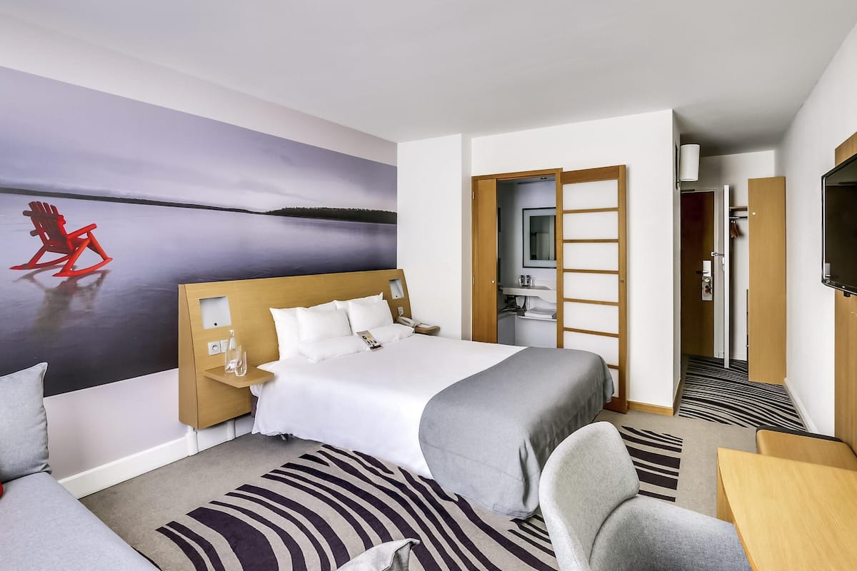 Habitación de hotel con cama, escritorio, silla, televisor y baño visible a través de una puerta abierta.
