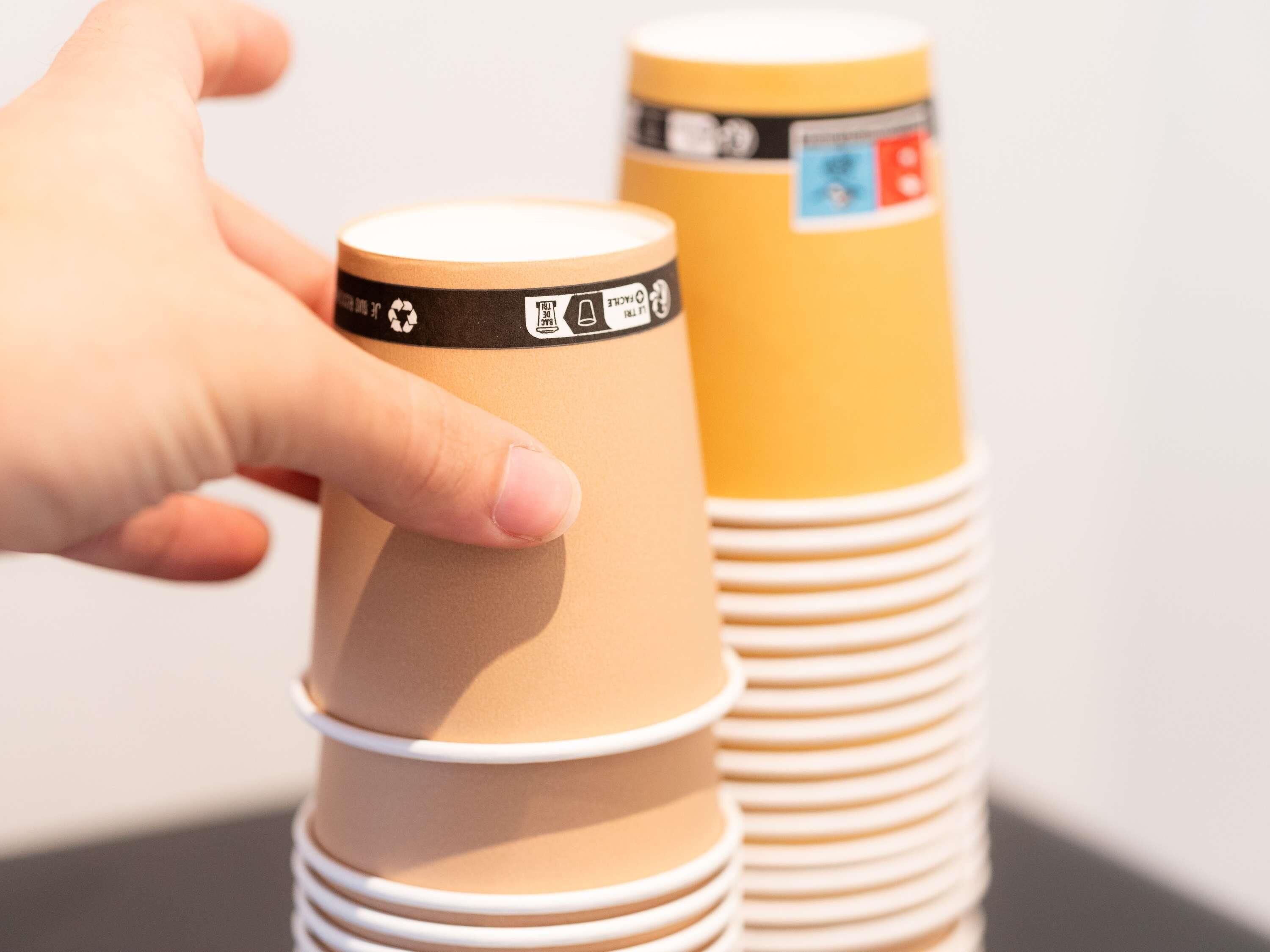 A hand holding a stack of paper cups with recycling symbols and a label.