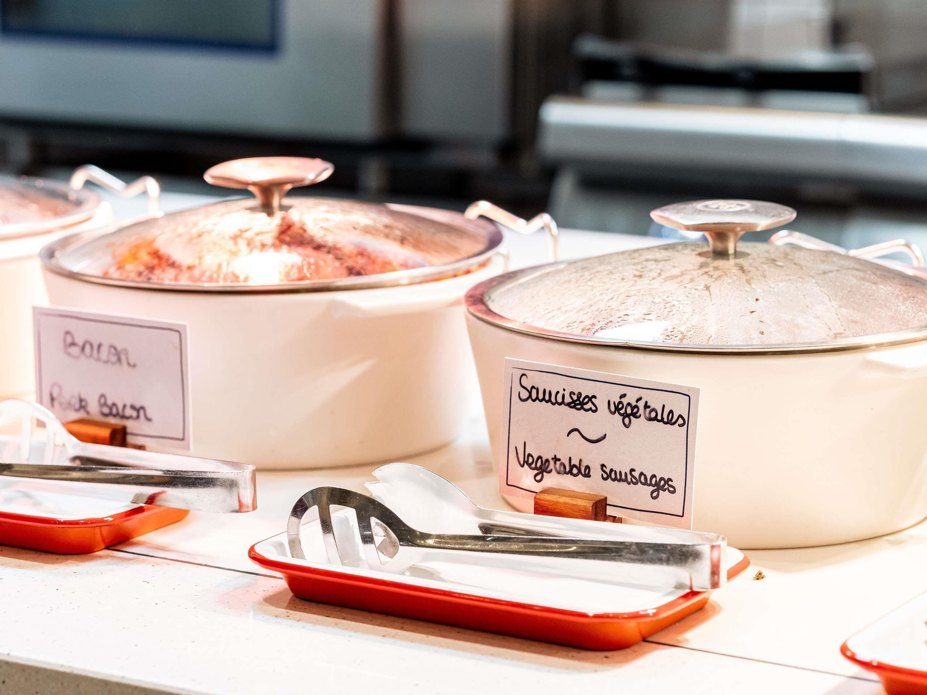 Buffet amb recipients etiquetats d'aliments, incloent 'Bacon' i 'Salsitxes vegetals'.