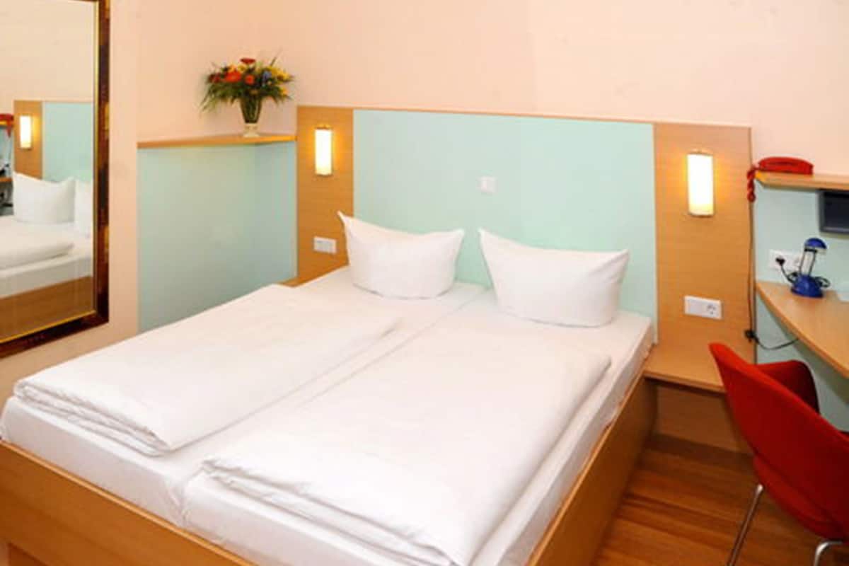 A double bed with white linens, a wooden headboard, a small desk with a red chair, and a wall-mounted lamp.