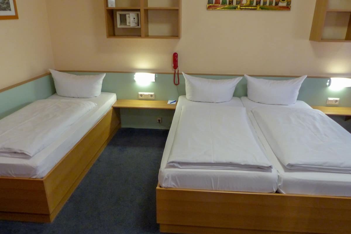 A hotel room with two single beds and a larger double bed, wooden headboards, a small bedside table with a red telephone, and a wall-mounted shelf with a framed picture.