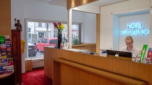 A hotel reception desk with a receptionist, a neon sign reading "HOTEL SACHSENHO", and a view of the street outside. - Hotel Sachsenhof Berlin (Berlin)