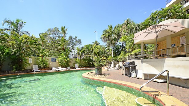 A clear swimming pool with a tiled edge, surrounded by lounge chairs and a covered seating area with a railing. - Marrakai Apartments (Darwin)