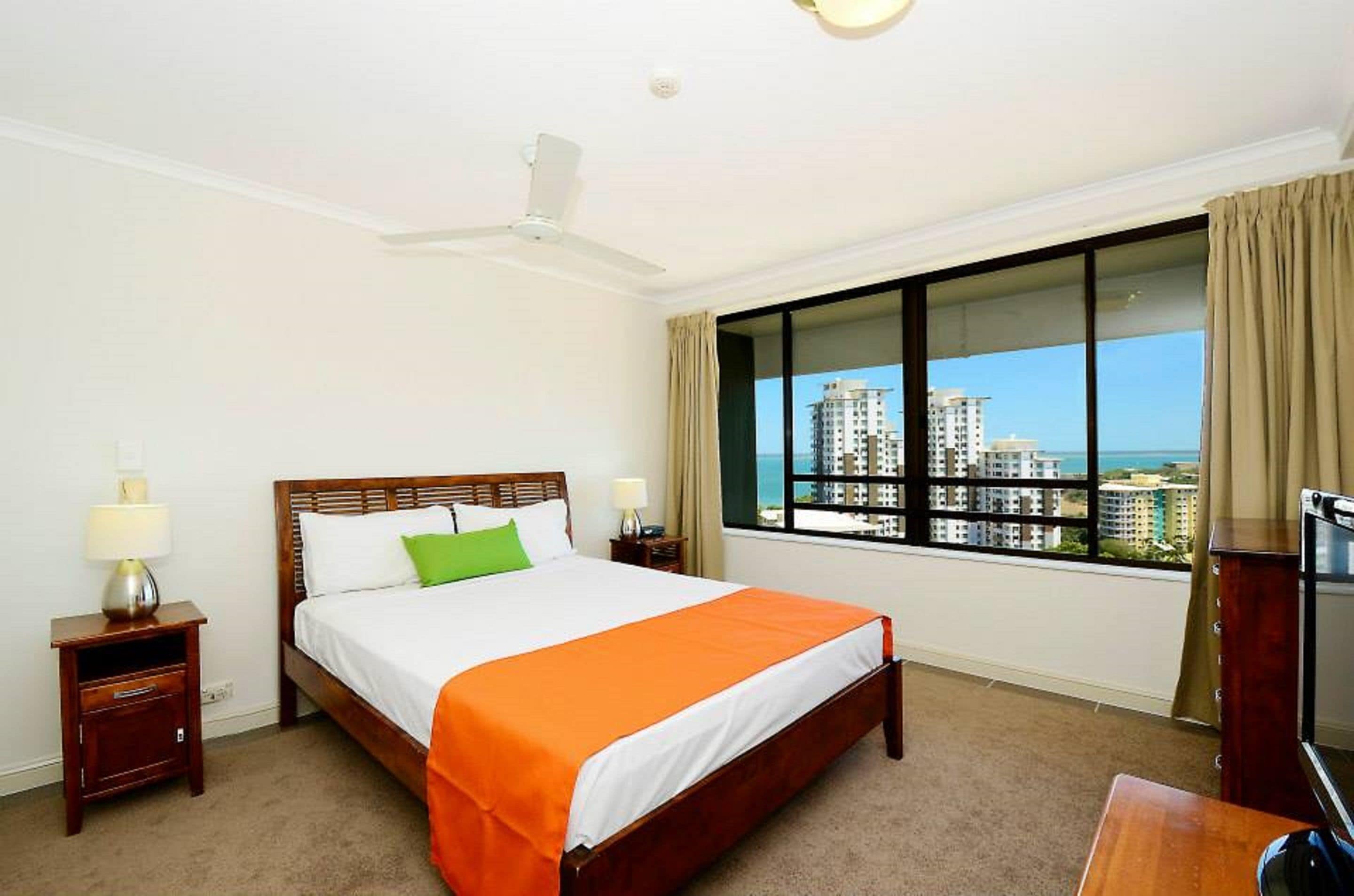 A hotel room with a large bed, two bedside tables, a ceiling fan, a TV, and a window with a city view.