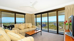 A spacious balcony with a sofa and a coffee table, large windows offering a view of the cityscape and construction cranes. - Marrakai Apartments (Darwin)