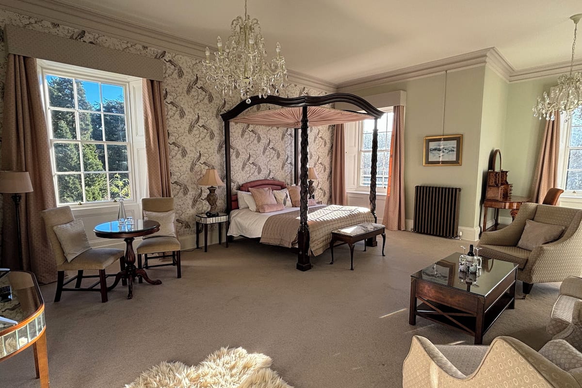 Een ruime slaapkamer met een hemelbed, een zithoek met een bank en fauteuils, een kroonluchter, en uitzicht op het omliggende landschap door grote ramen.