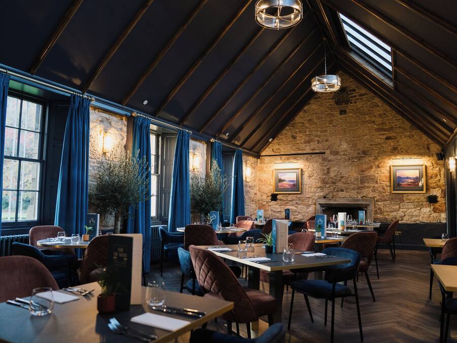 A restaurant with high ceilings, stone walls, and large windows with blue curtains. Tables are set with white tablecloths and chairs.