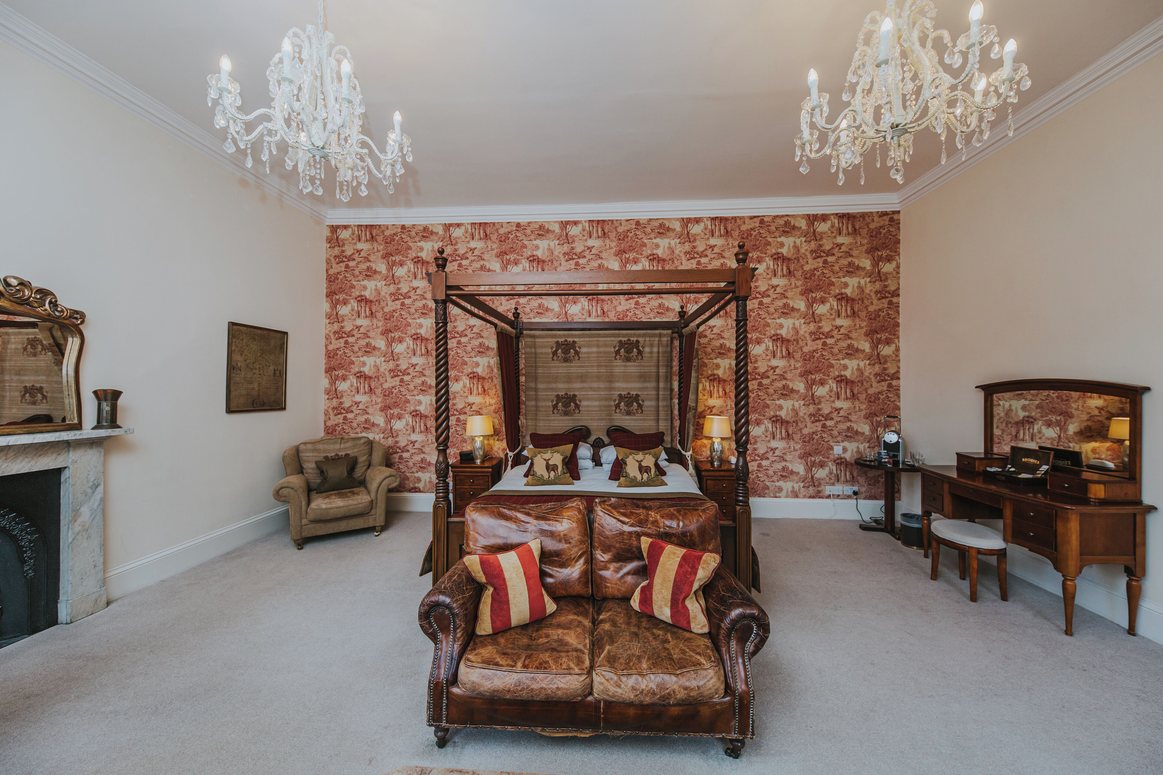 A bedroom with a four-poster bed, a fireplace, a chaise lounge, a desk, and a chandelier.