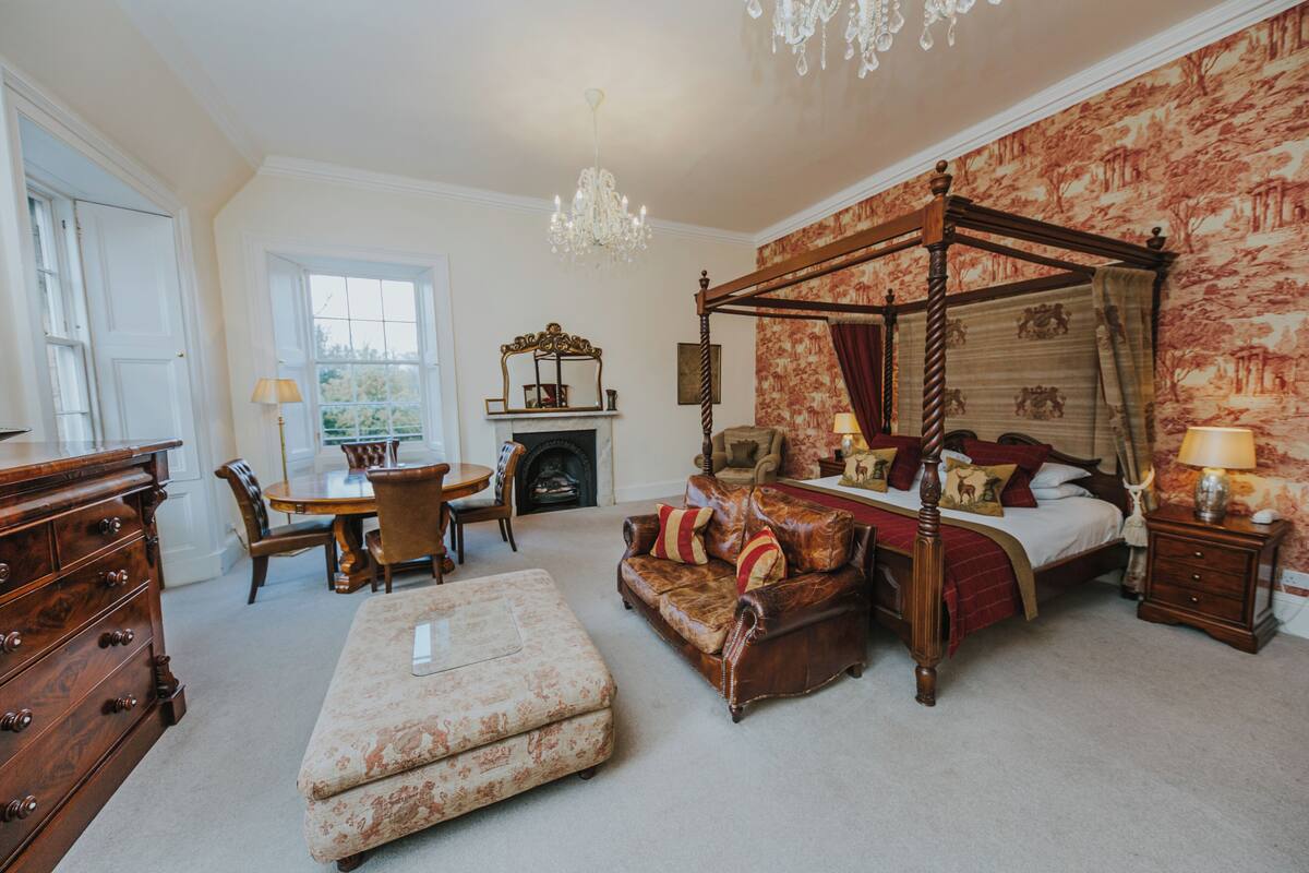 Een traditionele slaapkamer met een hemelbed, een open haard, een kroonluchter en een zithoek met een tafel en stoelen.