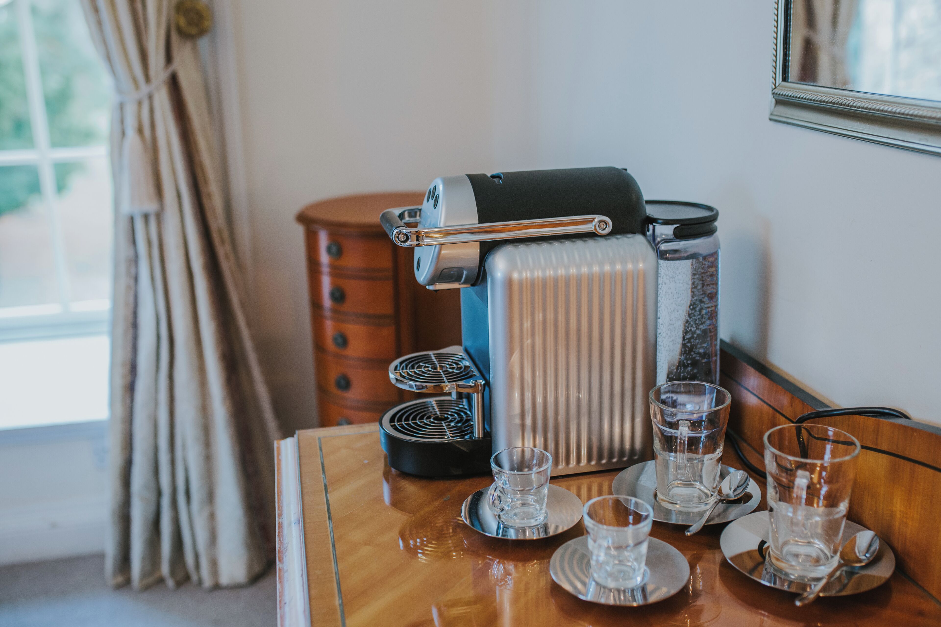 A coffee machine on a wooden table with four glasses and saucers, a drawer, and a window with curtains.