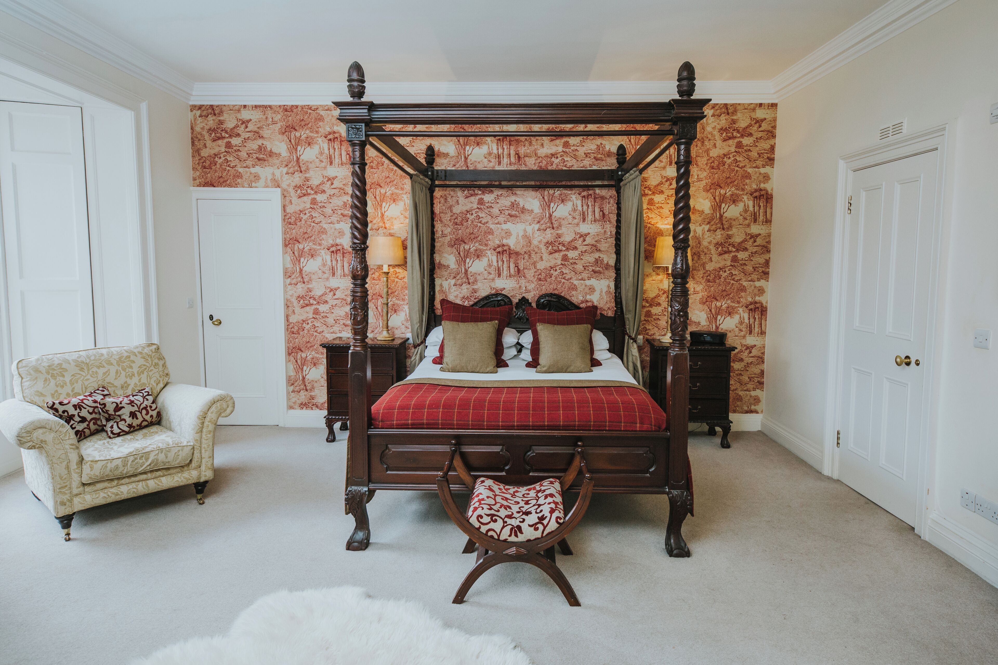 A four-poster bed with a red and white checkered bedspread, flanked by two nightstands with lamps, a chair, and a bench in a room with patterned wallpaper and carpeted flooring.