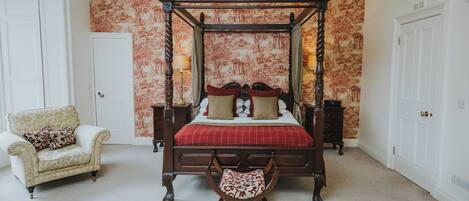 A four-poster bed with a red and white checkered bedspread, flanked by two nightstands with lamps, a chair, and a bench in a room with patterned wallpaper and carpeted flooring.