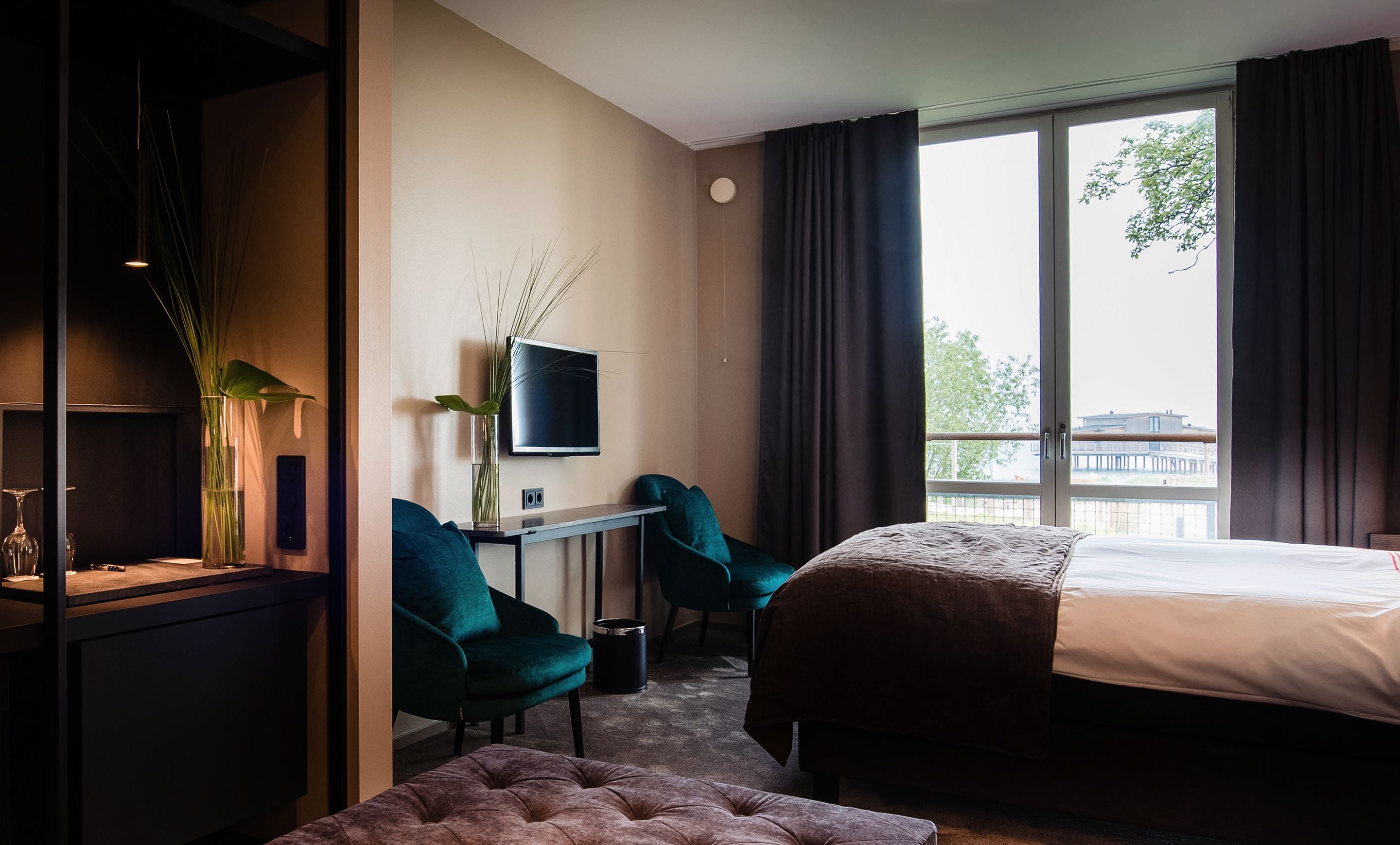 Ett hotellrum med en säng, ett skrivbord med en tv, två stolar, en växt och ett fönster med utsikt över en byggnad och träd.