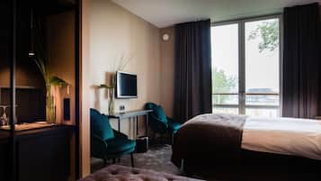 Ett hotellrum med en säng, ett skrivbord med en tv, två stolar, en växt och ett fönster med utsikt över en byggnad och träd.