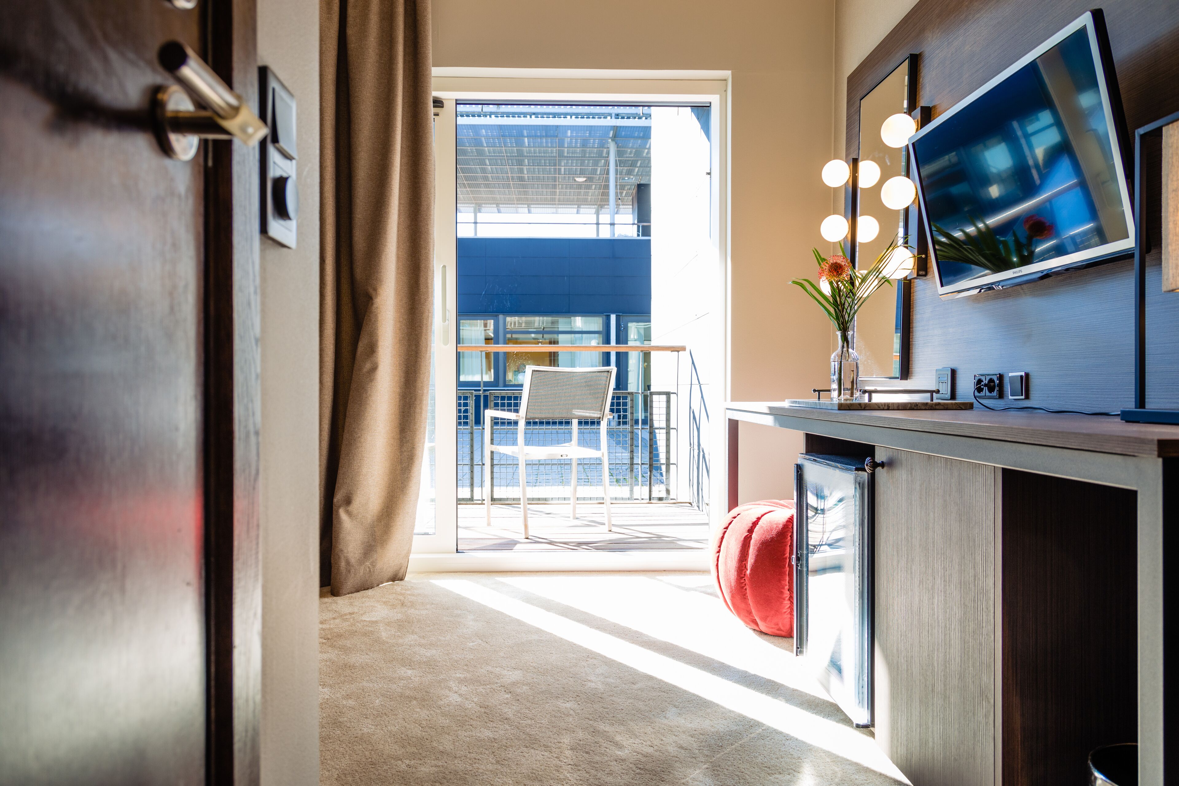 A modern kitchen with a built-in TV, a red ottoman, and a view of a balcony with a chair and table.