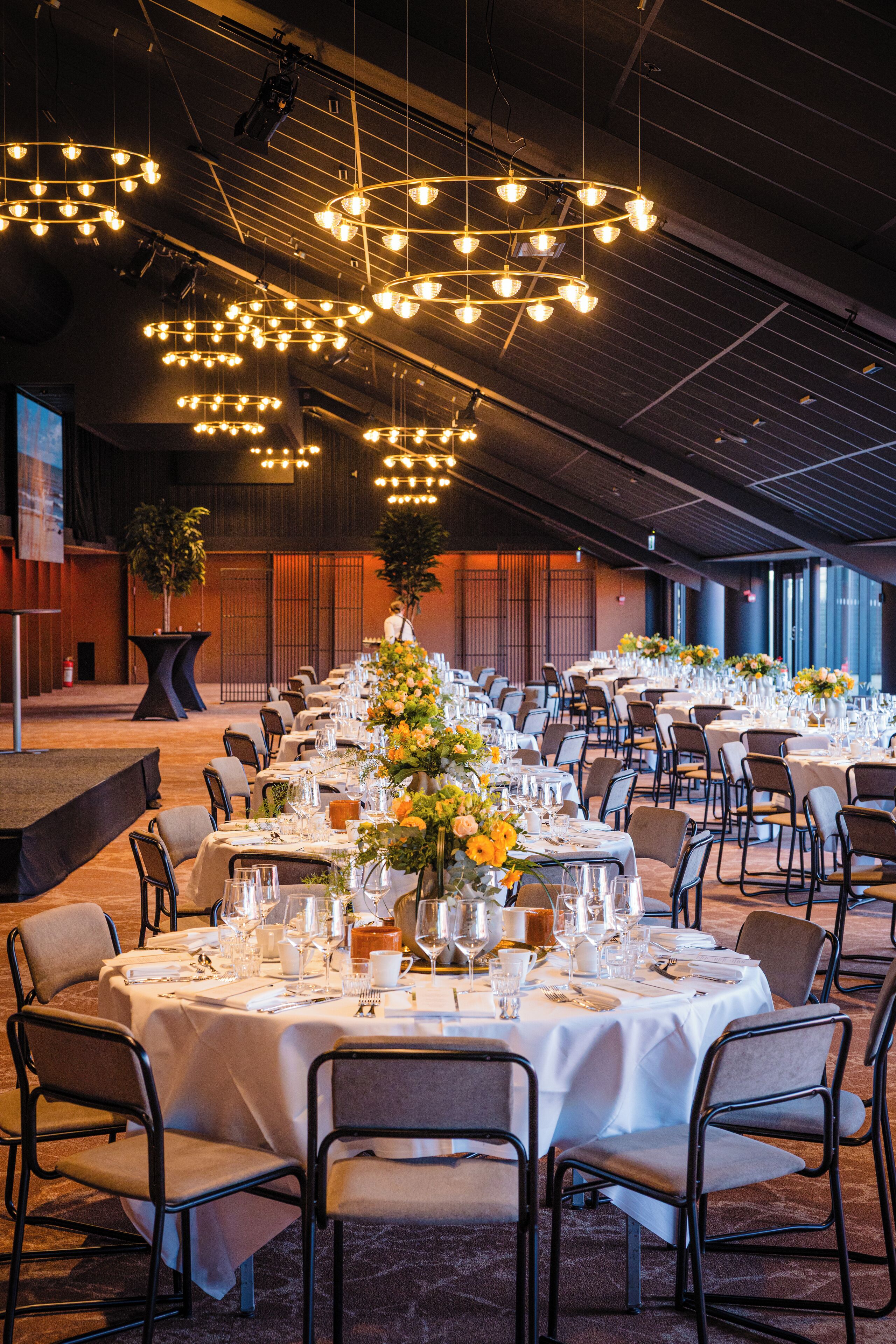 A banquet hall with round tables set for dining, overhead lighting, and a stage area.