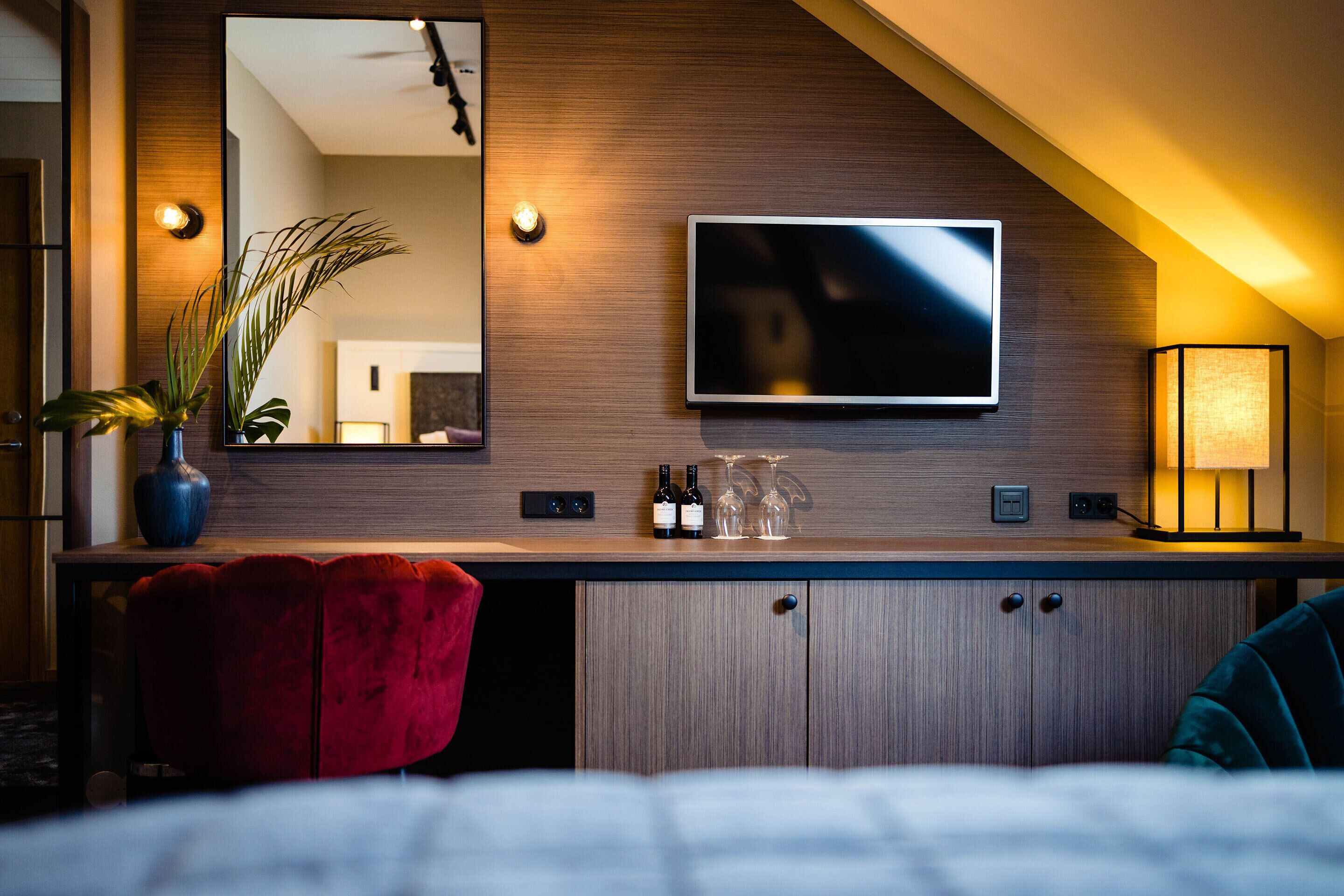 A modern hotel room with a wooden wall, a flat-screen TV, a red chair, a small table with wine bottles and glasses, and a bedside lamp.