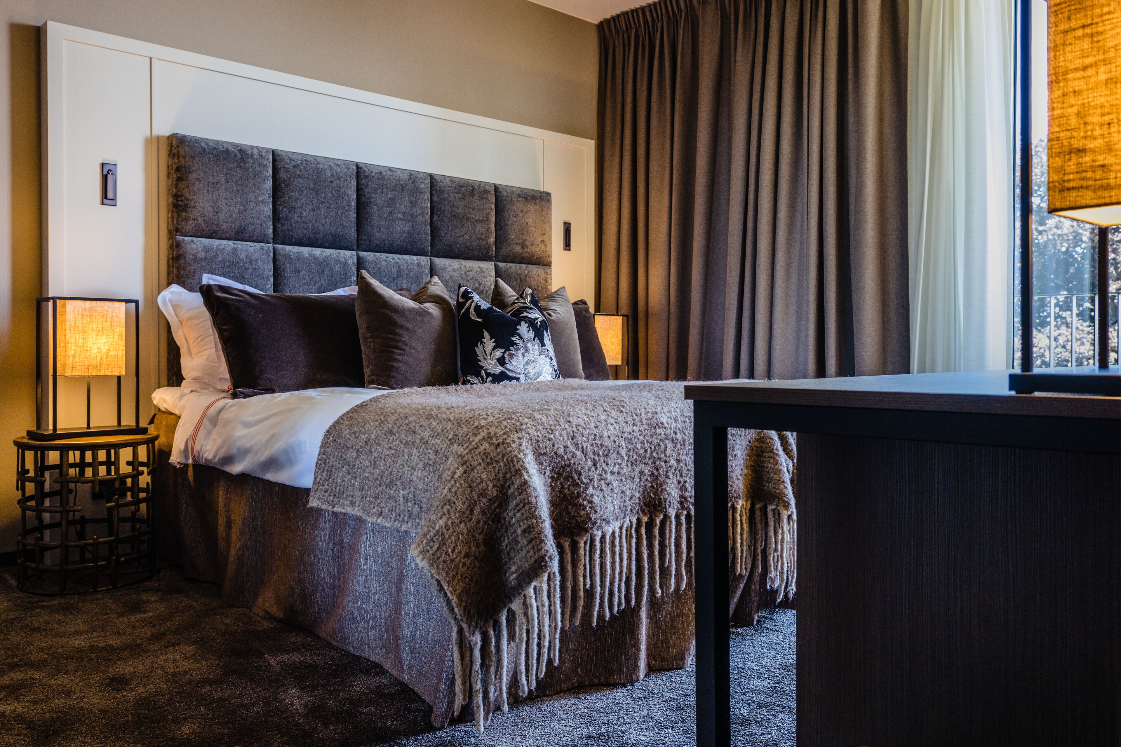 A bedroom with a large bed, a bedside table, a lamp, and a view of the outdoors through a window.