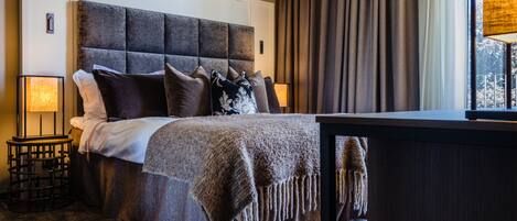 A bedroom with a large bed, a bedside table, a lamp, and a view of the outdoors through a window.