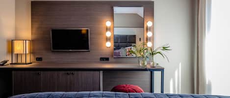 A hotel room with a bed, a TV, a vanity with lights, and a plant.