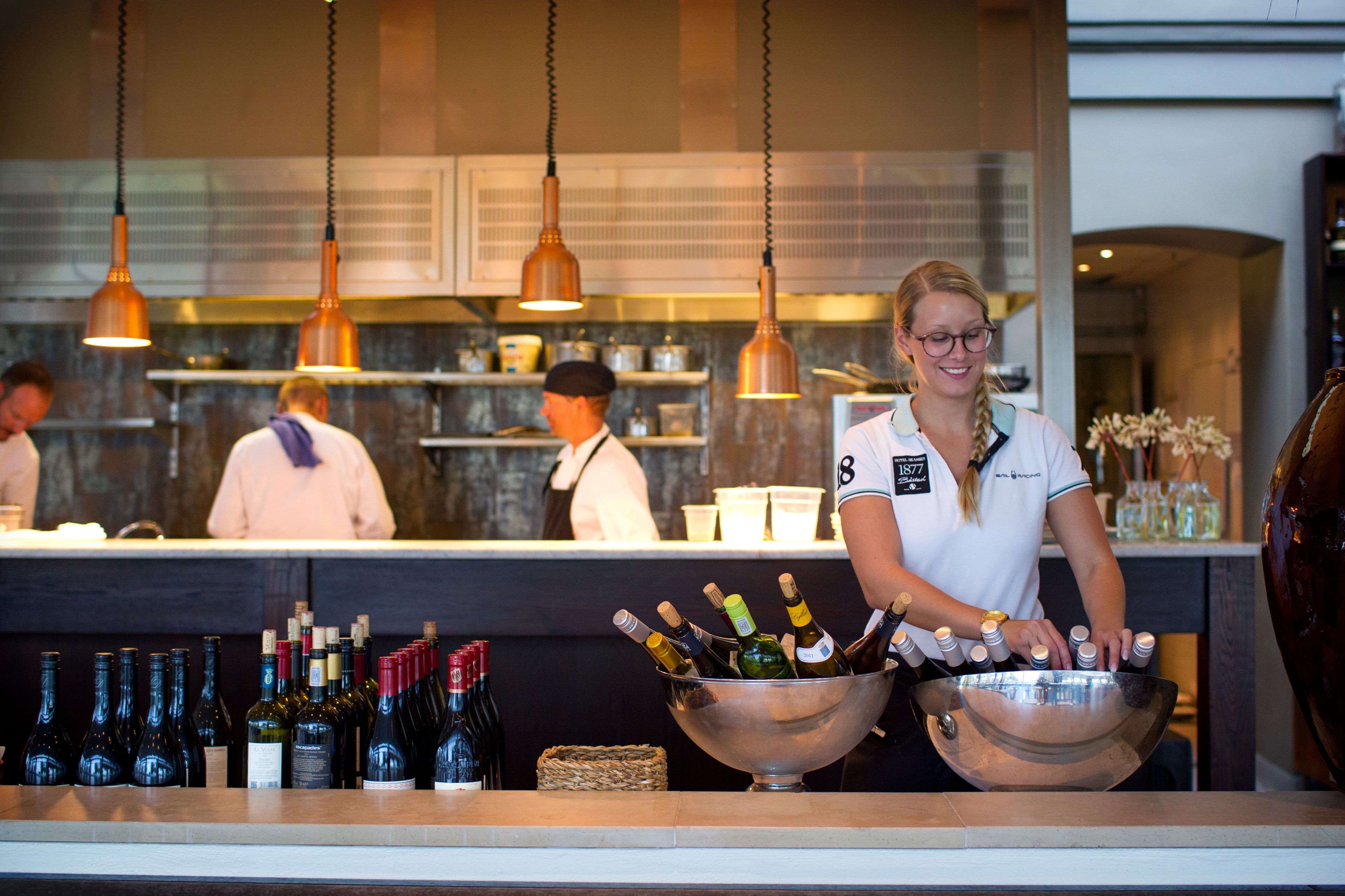 A bar area with a bartender preparing drinks, a counter with bottles, and a kitchen in the background.