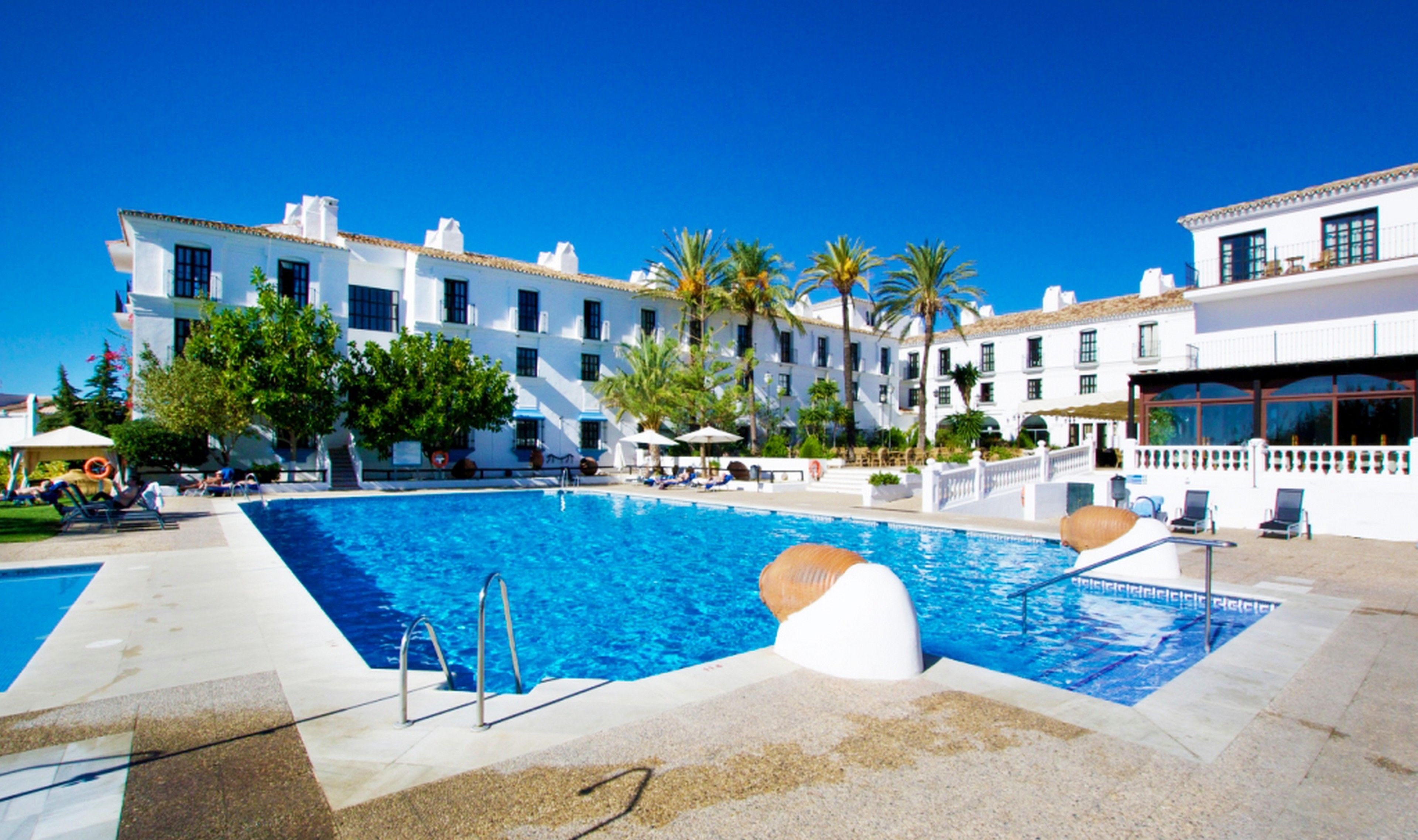 A swimming pool with a clear blue water, surrounded by white buildings with multiple windows and balconies, and palm trees.
