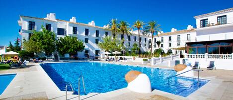 A swimming pool with a clear blue water, surrounded by white buildings with multiple windows and balconies, and palm trees.