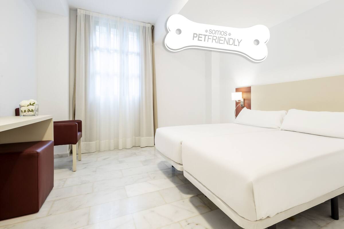 Quarto de hotel com uma cama grande, uma mesa e uma cadeira. Há um aviso na parede que diz "Somos PETFRIENDLY".