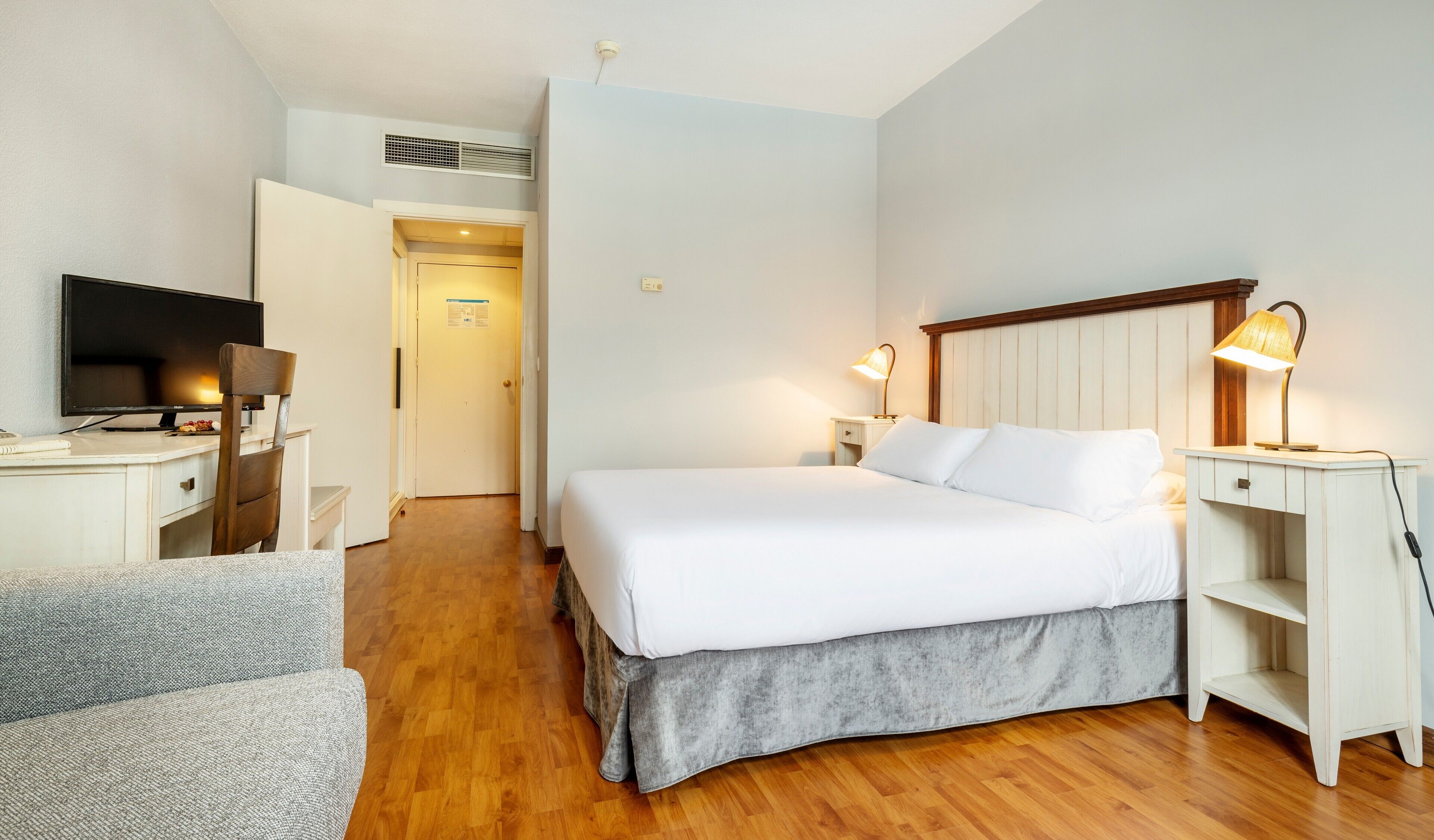 A hotel room with a bed, bedside tables, a desk with a television, a chair, and a lamp.