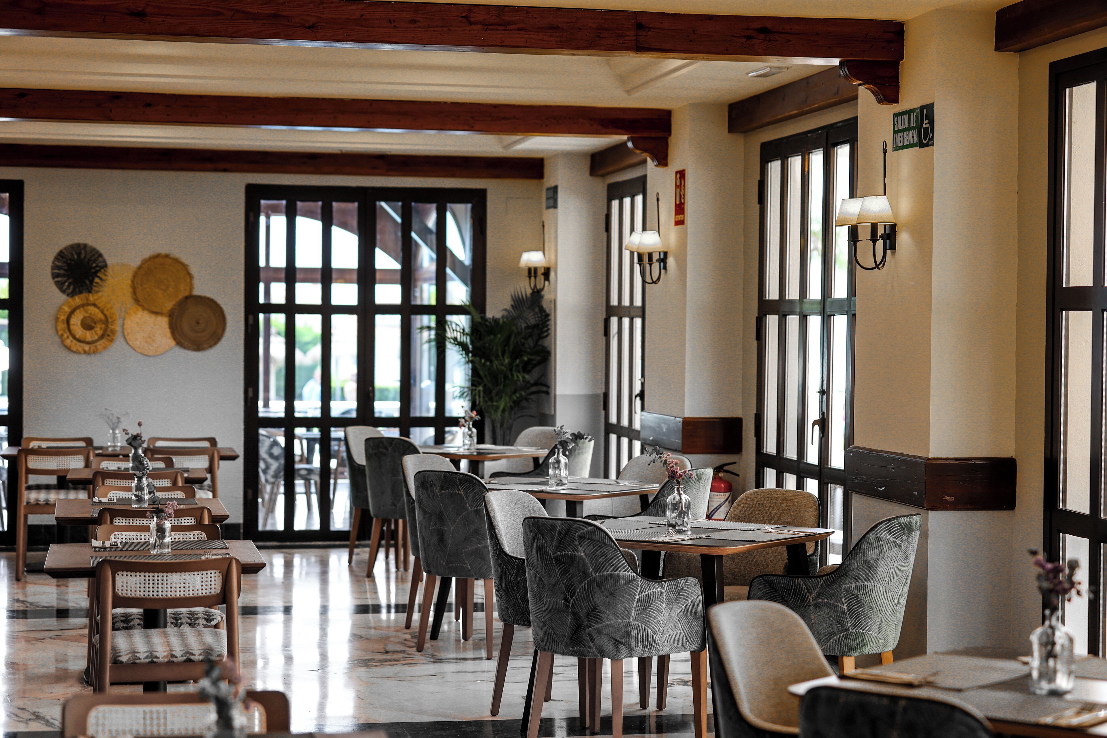 Un restaurant au décor moderne, avec un mélange de chaises en bois et de chaises rembourrées, des tables agrémentées de centres de table et de grandes fenêtres laissant entrer la lumière naturelle.