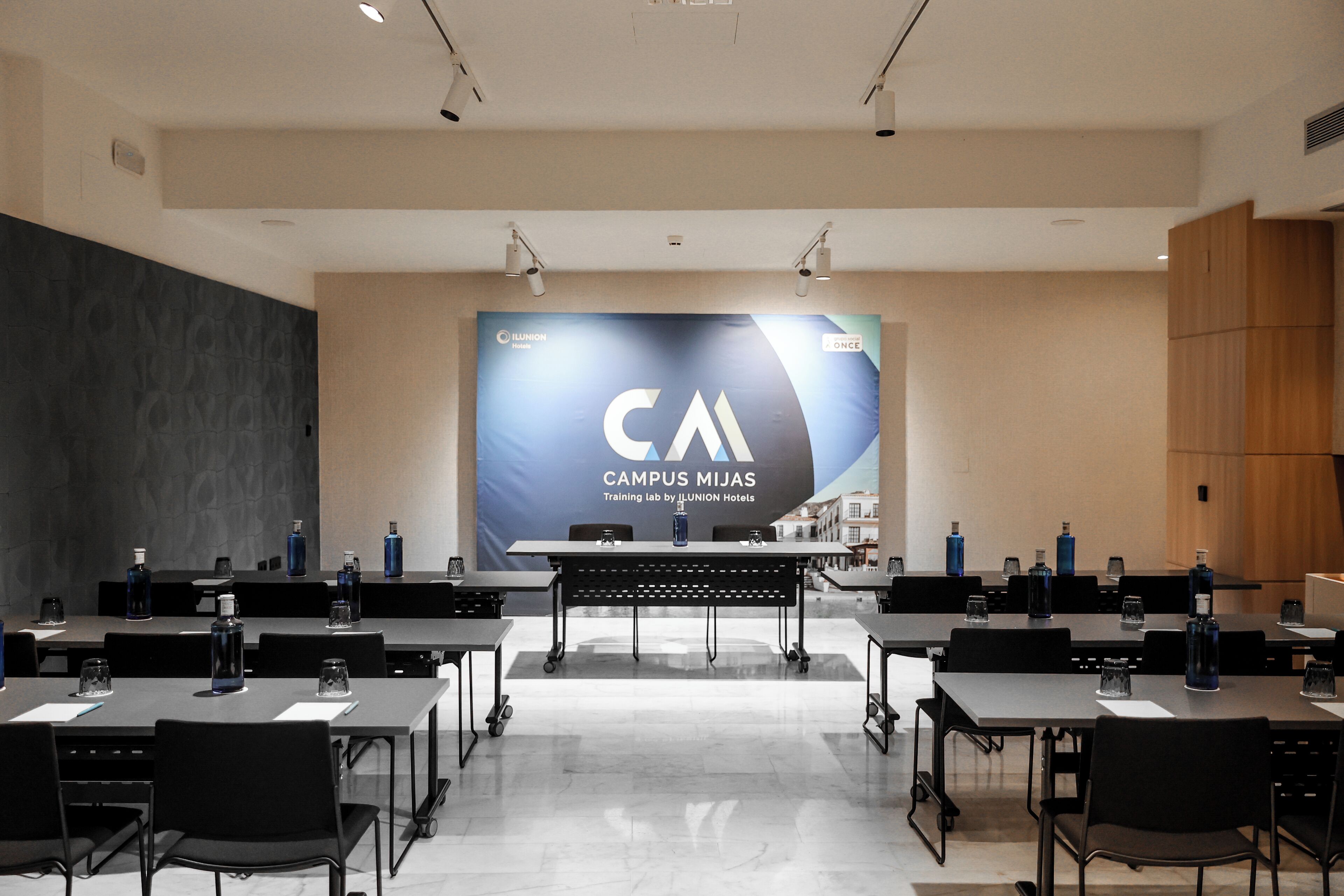 A conference room with multiple tables, chairs, and a large screen displaying 'CAMPUS MIJAS'.