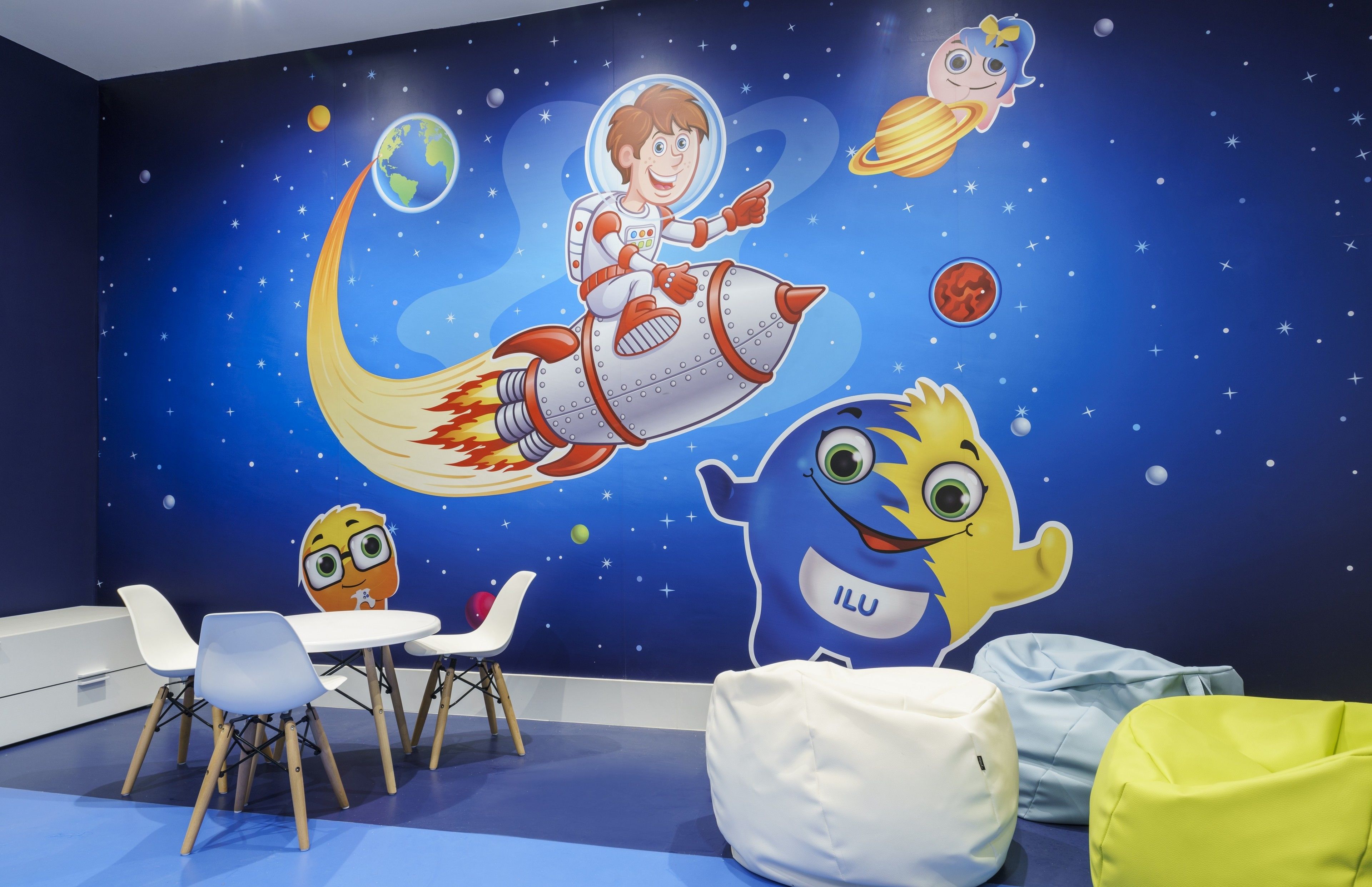 Ein Kinderspielzimmer mit Wandmalerei zum Thema Weltraum, weißen und blauen Stühlen sowie Sitzsäcken.