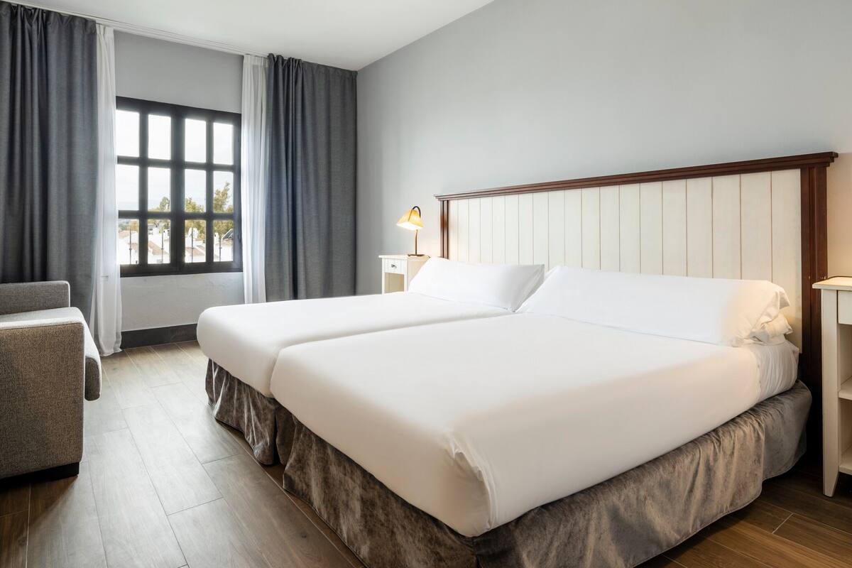 Quarto de hotel com cama de casal, uma cadeira, mesa de cabeceira com abajur e janela com cortinas.