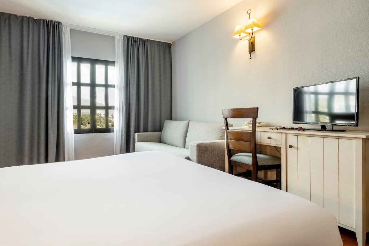 Quarto de hotel com uma cama grande, uma mesa de trabalho com uma TV, uma cadeira e vista pela janela.