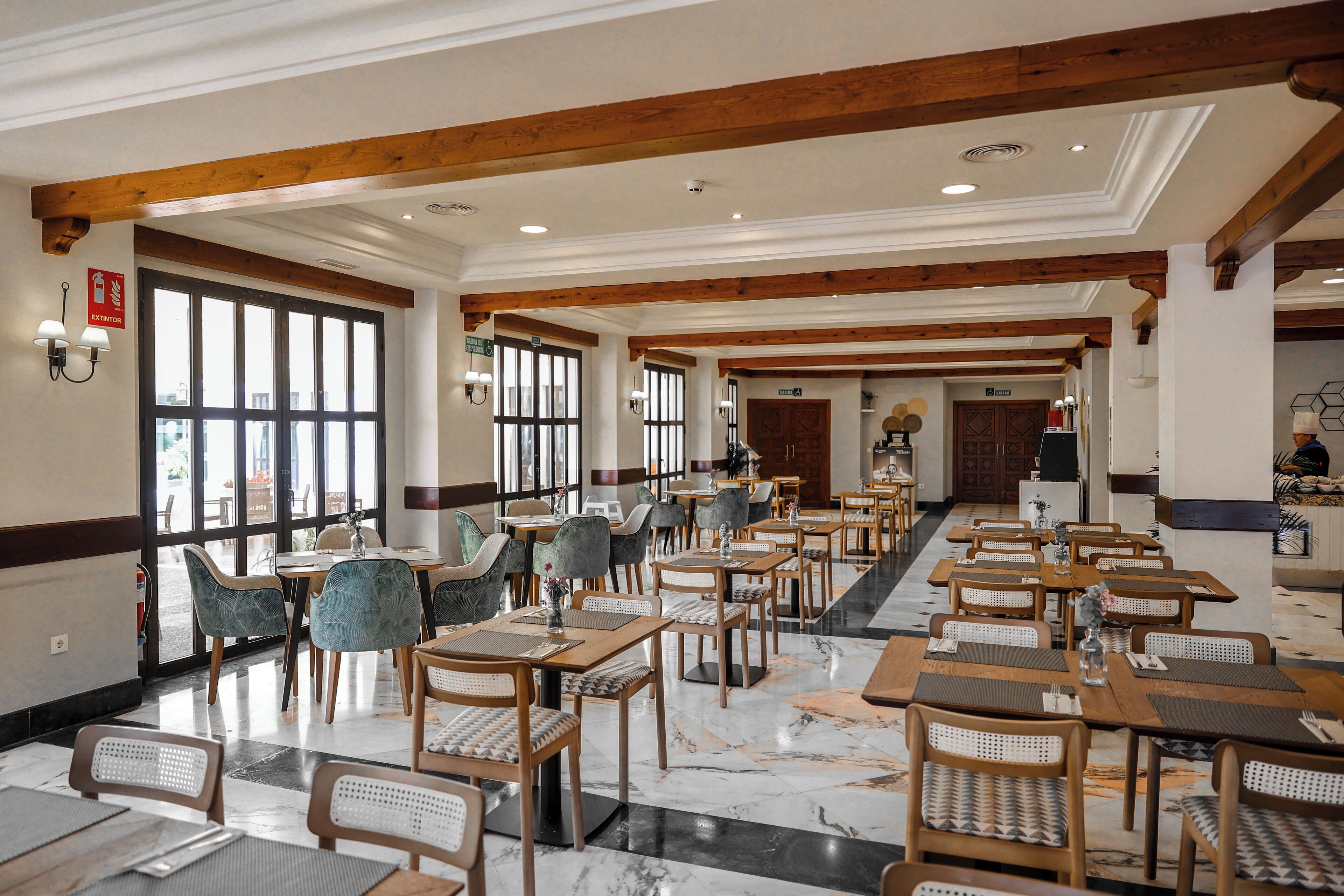 A spacious dining area with wooden tables and chairs, large windows, and a high ceiling with exposed beams.